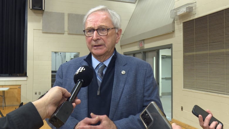 A man with grey hair and glasses, wearing a collared shirt, tie and blazer, speaks into several microphones and recorders being held by reporters.