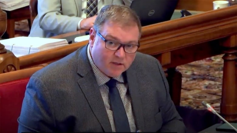 A man with short brown hair and glasses, wearing a suit, seated at a table, speaking into a microphone.