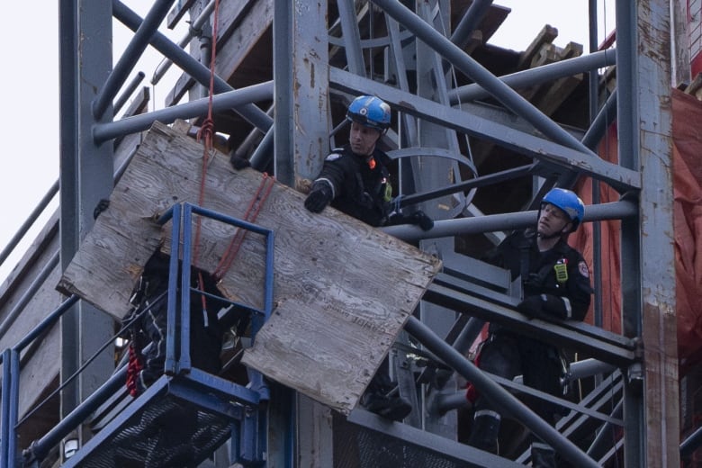 Two men wearing blue helmets are dwarfed by the scaffolding they're on. One man grips a large piece of wood with one hand.