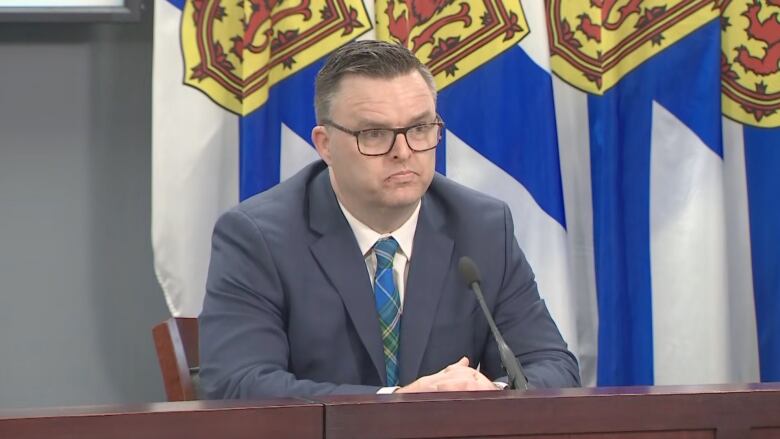 A man in a suit is seated in front of Nova Scotia flags.