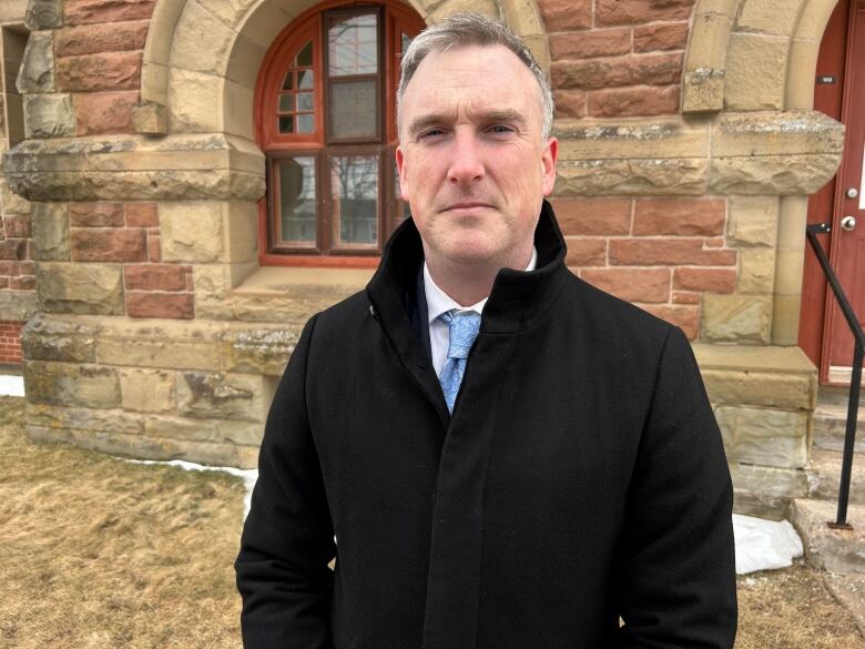 A man with close-cropped light hair and a black overcoat over a shirt and blue tie.