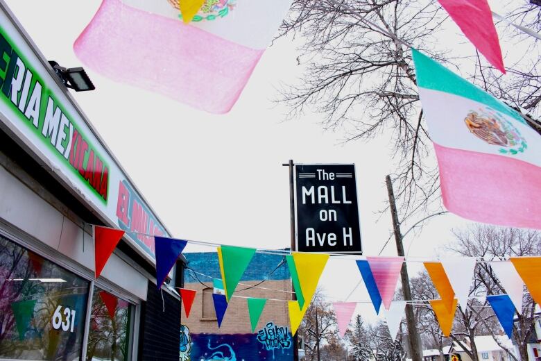 Multi-coloured triangular flags and the Mexican flag fly outside of a business. A large street sign reads 'The Mall on Ave H.'