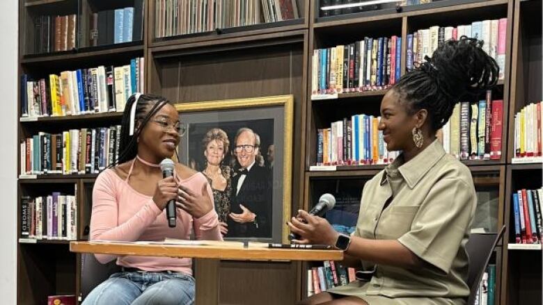 A black woman in glasses and a pink top holds a mic, speaking to another black woman with long tied up hair in a green dress sitting across a table from one another.