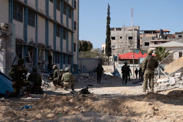 A group of soldiers, some crouching and some standing, between a damaged concrete building and piles of concrete chunks and debris.