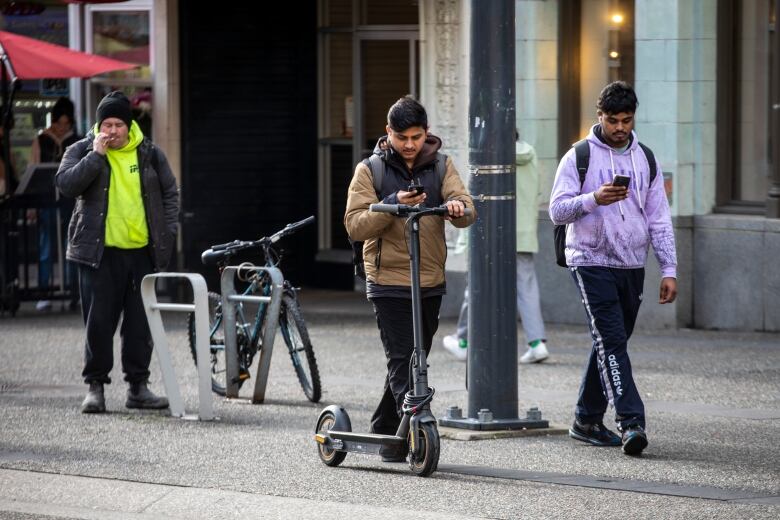 A man in a khaki-coloured jacket is seen walking with an e-scooter. Behind him, to his left, is a man in a purple hooded sweatshirt walking while looking on his phone. Further behind, a man in a neon-yellow-coloured sweatshirt and a toque is seen smoking a cigarette.