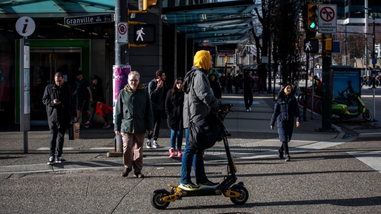 A person with a yellow hooded sweatshirt and an olive jacket is seen riding an e-scooter on a busy street.