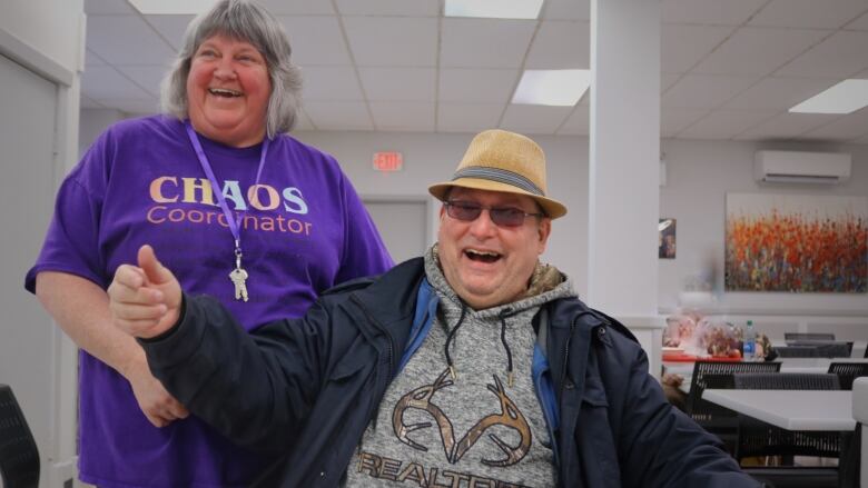  A smiling older woman in a purple shirt and a laughing man in a black windbreaker joke around in a cafeteria