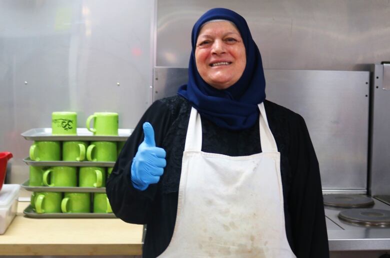 A woman in a hijab gives a thumbs up sign in front of a stack of bright green coffee mugs. 