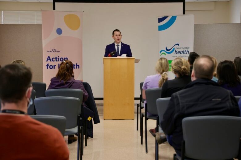 a white man is seen from some distance away while standing a a podium and speaking to several seated people.