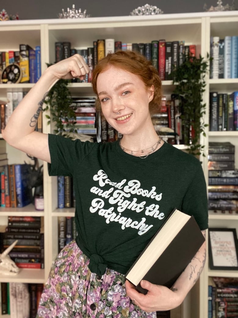 A young woman flexes a bicep and holds a book standing in front of a full bookcase. Her T-shirt says 