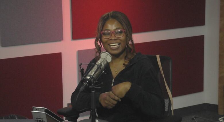 woman wearing glasses and a black jacket smiling and sitting in front of a microphone