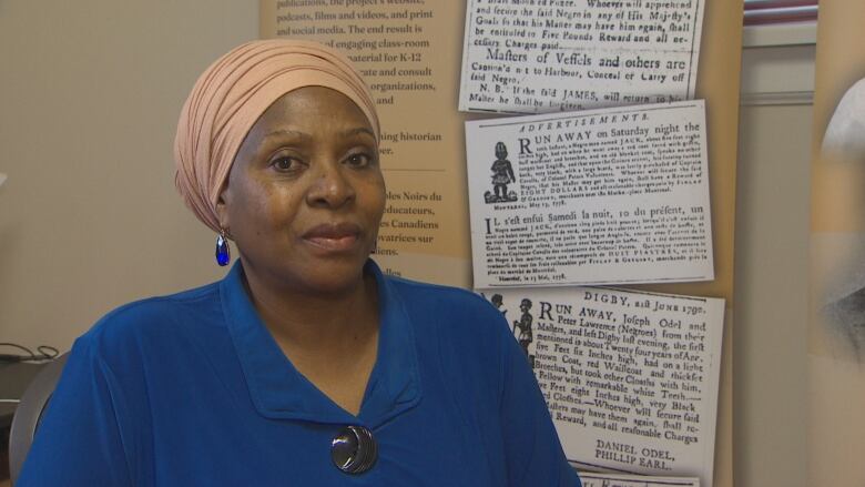 A woman in a blue shirt, drop earrings and a blush-coloured headwrap sits in an office in front of sign depicting archival newspaper clippings.