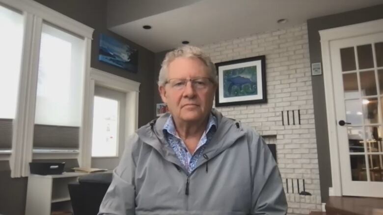 A man with grey hair in a grey windbreaker sits in an office facing the camera.