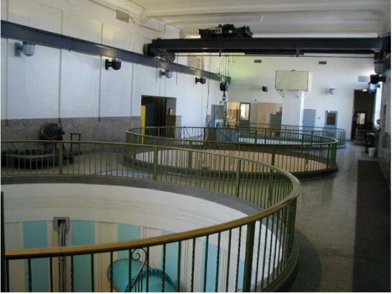 A photo shows the inside of a large industrial building. There are three large circular holes in the floor with railing around them.