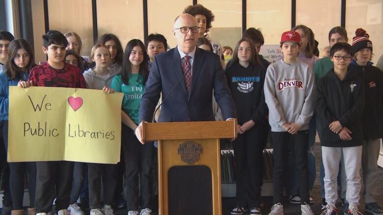 A man is pictured standing at a podium, with a group of children standing behind him. One of them holds a sign that says 