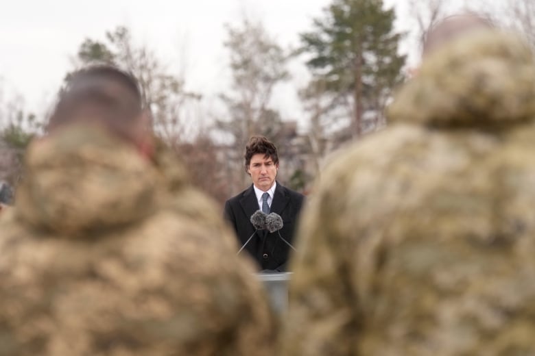 A man speaks at a podium, with two soldiers blurred in the foreground.