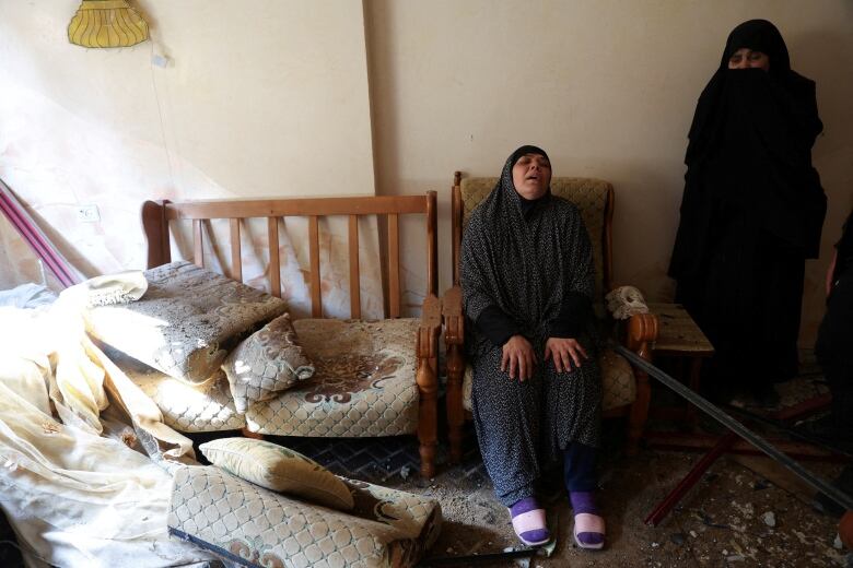 Two people, one sitting in a chair and another standing beside them, react in despair inside a home with belonging and rubble strewn on the ground.