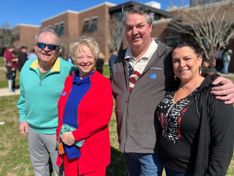 Group photo of elderly man and woman, he in sunglasses, she in a bright red coat, next to a middle-aged couple. In the background: a long line outside a brick school that was a polling station