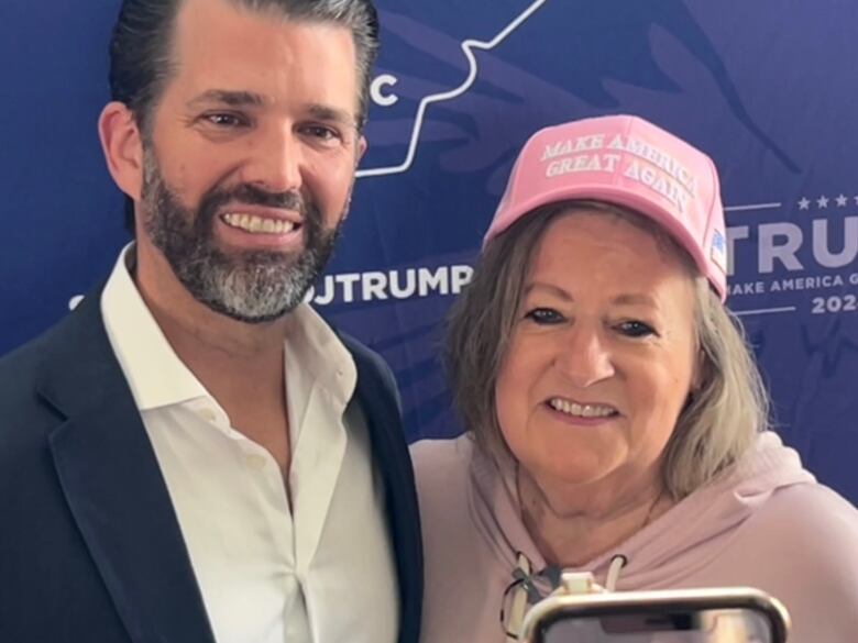 Bearded man in shirt and sport coat, Donald Trump Jr., poses for photo with woman in pink Make America Great Again cap. 