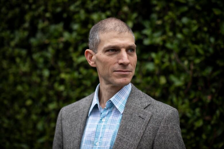 A white man wearing a grey jacket and a blue checked shirt poses against a backdrop of a hedge.