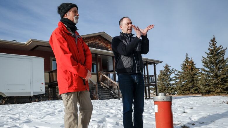 Two men stand on a snow covered lawn with a house behind them and a red tube protruding from the ground just in front of them. 