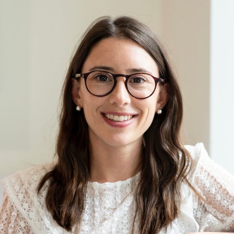 A Caucasian woman with brown hair and glasses stands indoors. She is wearing a white shirt.