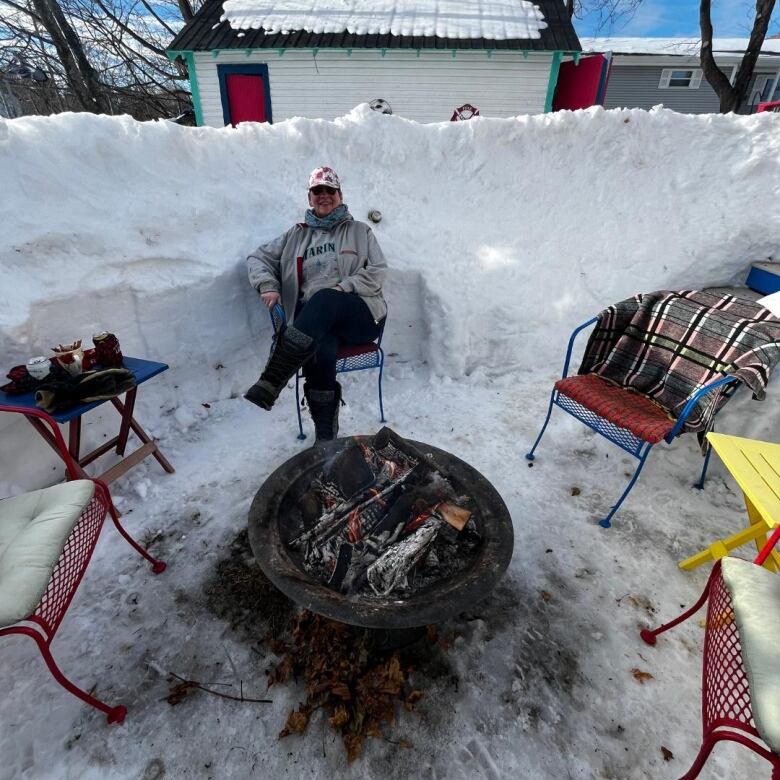 A woman dressed in a grey hoodie and white basketball cap sits in a lawn chair in front of an outdoor fireplace. Behind her is an enormous wall of snow.