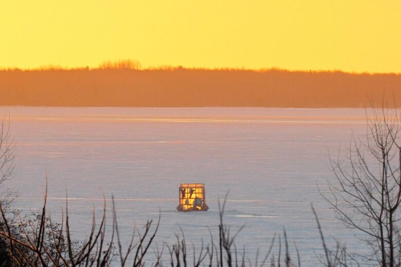 A small, illuminated square structure sits on a frozen body of water beneath a glowing, yellow sky.