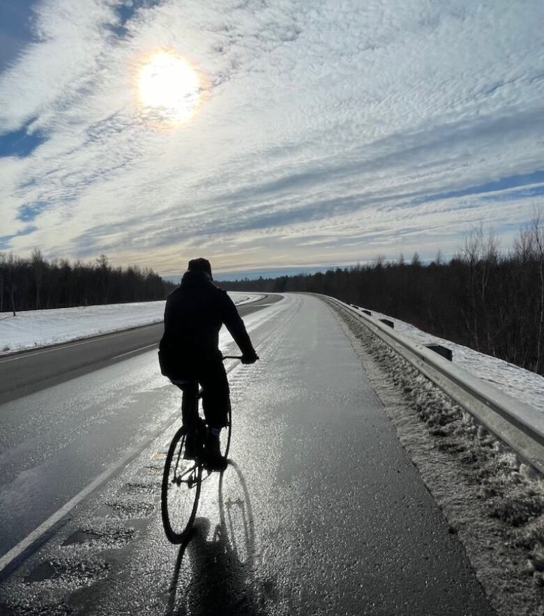 A person rides a bike down a road bordered by snow on each side. The sun glares down from high in the sky.