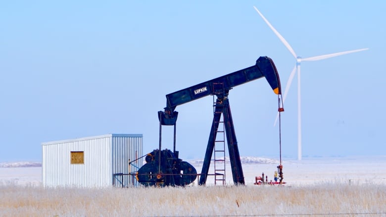 A wind turbine is pictured in the distance behind an oil pumpjack in a field.