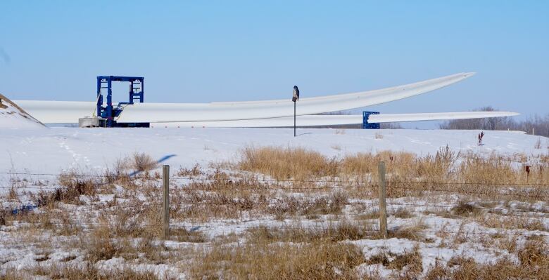 Two white wind turbine blades are on the ground in a snowy field.