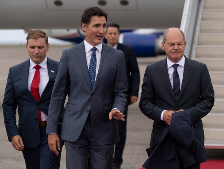 Three men wearing suits walk towards the camera with an aircraft in the background.