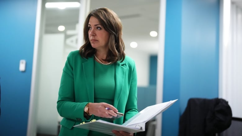 A woman wearing a green blazer with white pearls with brown hair is pictured in front of a blue wall.