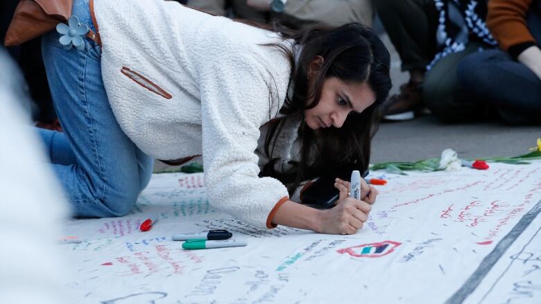 A woman writes with a marker on a large piece of paper while kneeling in what appears to be an outdoor setting.