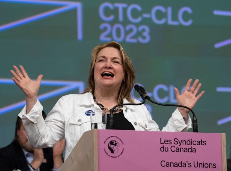 A woman in a white jacket gestures with her hands as she speaks into a microphone at a podium.