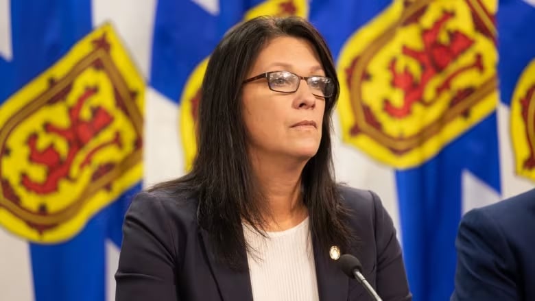 A woman with dark hair and glasses, wearing a blazer.