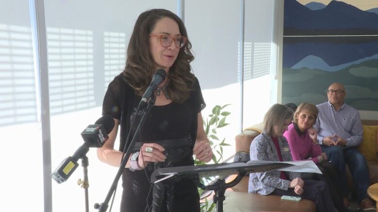 a woman with glasses stands at a podium in a small room. she speaks into the microphone while a group of people listen.