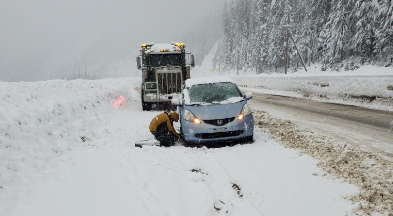 A person crouches beside a small, blue car on the side of a snowy highway with a transport truck parked behind it.