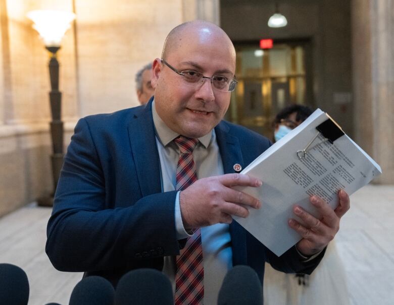 A bald man with glasses holds up a stack of papers, standing in front of a microphone