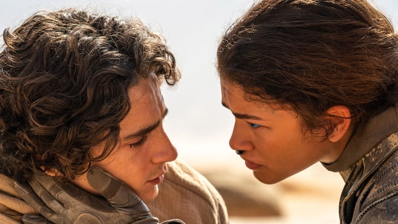 A man and woman stare longingly into each other's eyes. Behind them are desert dunes. 