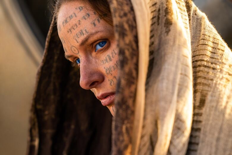 A woman with bright blue eyes peers out from under a hood. Her face is covered in tattooed letters of a non-english language.