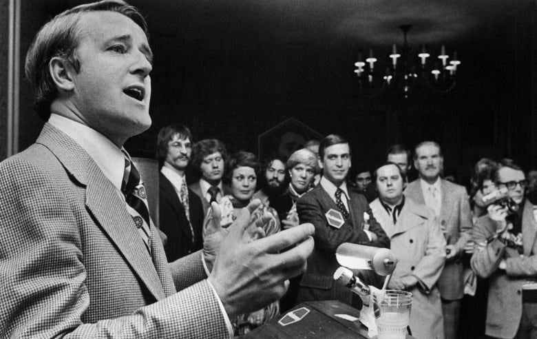 A black and white photo shows a man in a suit speaking to a crowd gathered around him.