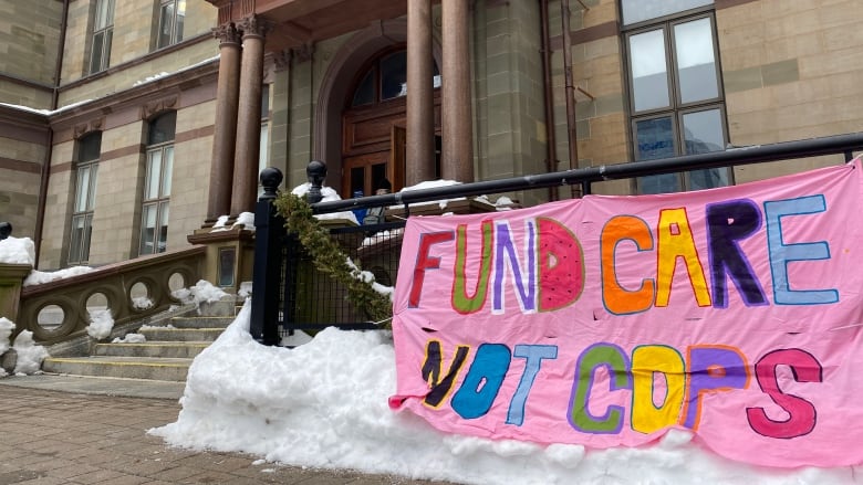 A pink banner with colourful writing hangs on the railing outside the brick front of City Hall. The sign reads 'fund care not cops'