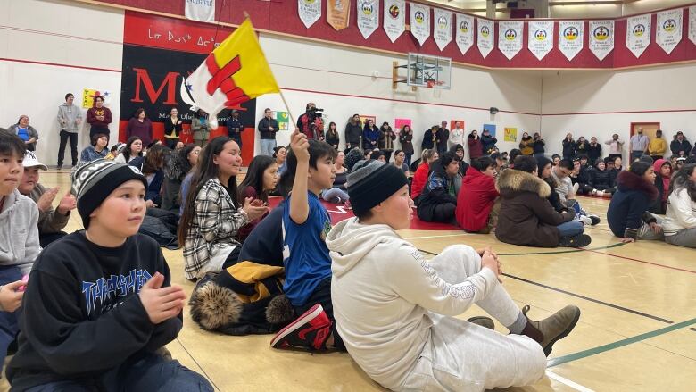 A crowd of people sit on the floor inside a school gymnasium, with one person holding up a Nunavut flag.