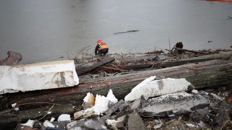 A man crouches at the shore in a rain coat and life preserver. In the foreground there are huge heaps of styrofoam and other junk.