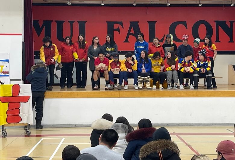 A group of people are seated on the stage inside a school gymnasium as other people on the gym floor look on.