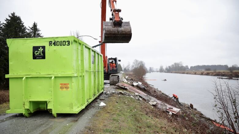 A large light green bin is in the foreground, orange excavator behind, garbage strewn around the bank down toward the river, where a man climbs around.