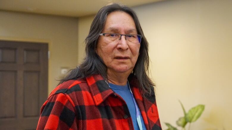 A person with long dark hair and a red plaid shirt stands in a room with a neutral expression on their face.