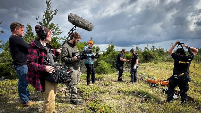 Four people with camera equipment are on the left side filming three firefighters who are working a hose. They are standing on a green piece of land with trees in the background and cloudy grey skies. 