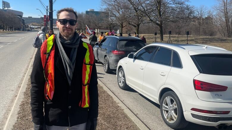 A young man in a high-visibility orange vest and sunglasses smiles lightly at the camera. A line of cars is behind him, along with a picket line. York University is far in the background.